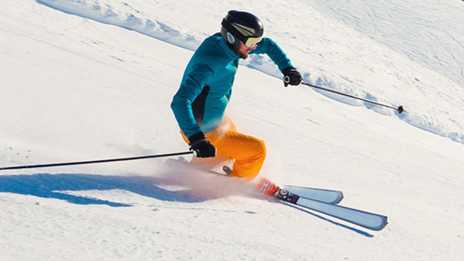 Un skieur en bleu et orange descend une pente de montagne enneigée et ensoleillée dans les Alpes.