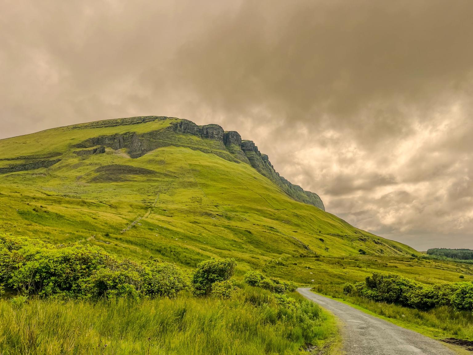 A green, grassy mountain with rocky ledges under a cloudy sky, with a curving road in the foreground.