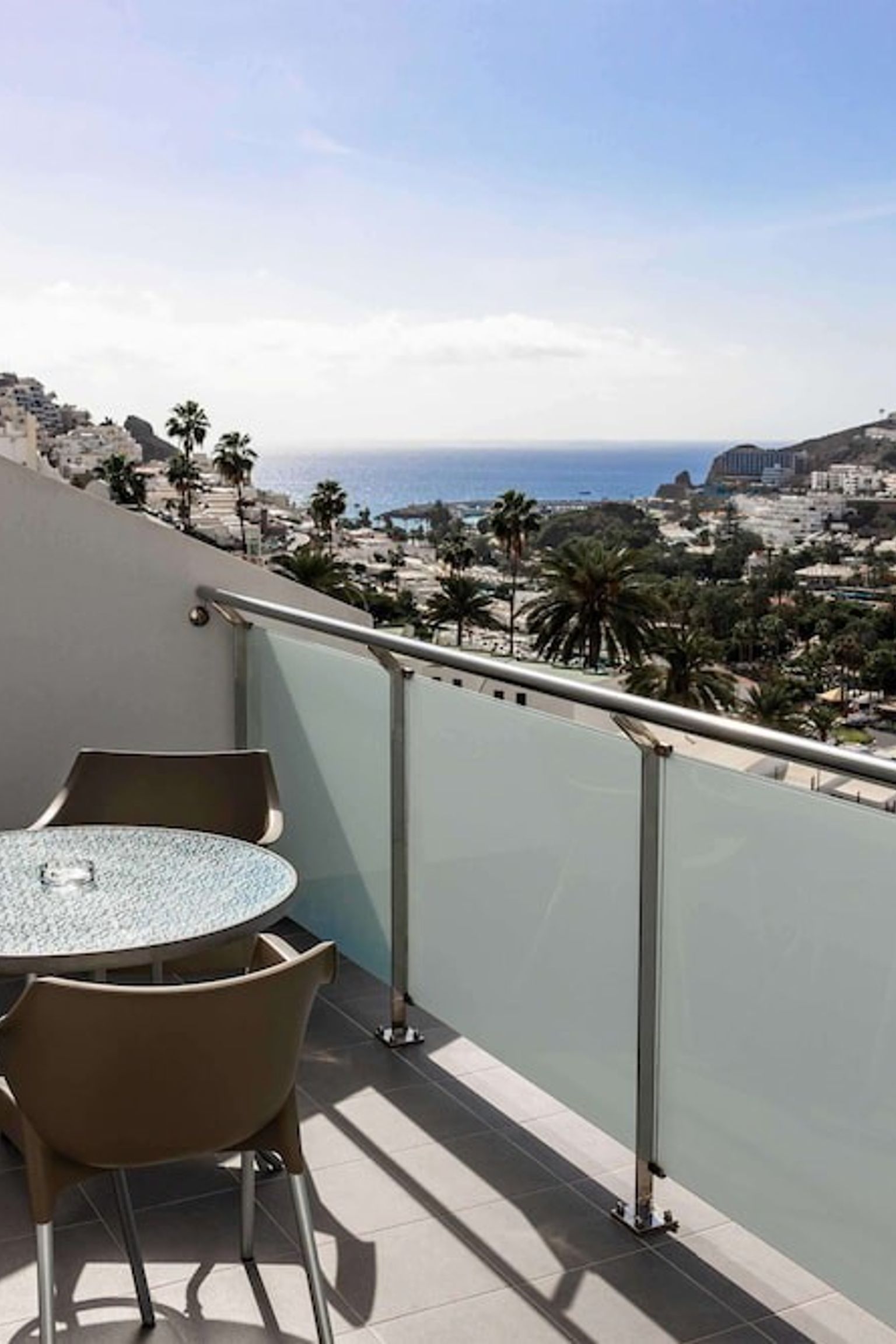En balkong med ett bord och stolar med utsikt över en stad på en sluttning, palmer och havet.