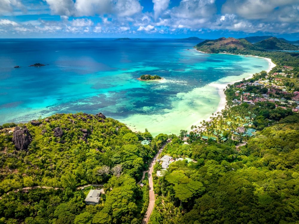 Flygfoto över en livlig tropisk kustlinje med en vit sandstrand, turkost vatten, frodiga gröna kullar och resorter. Praslin Island, Seychellerna.