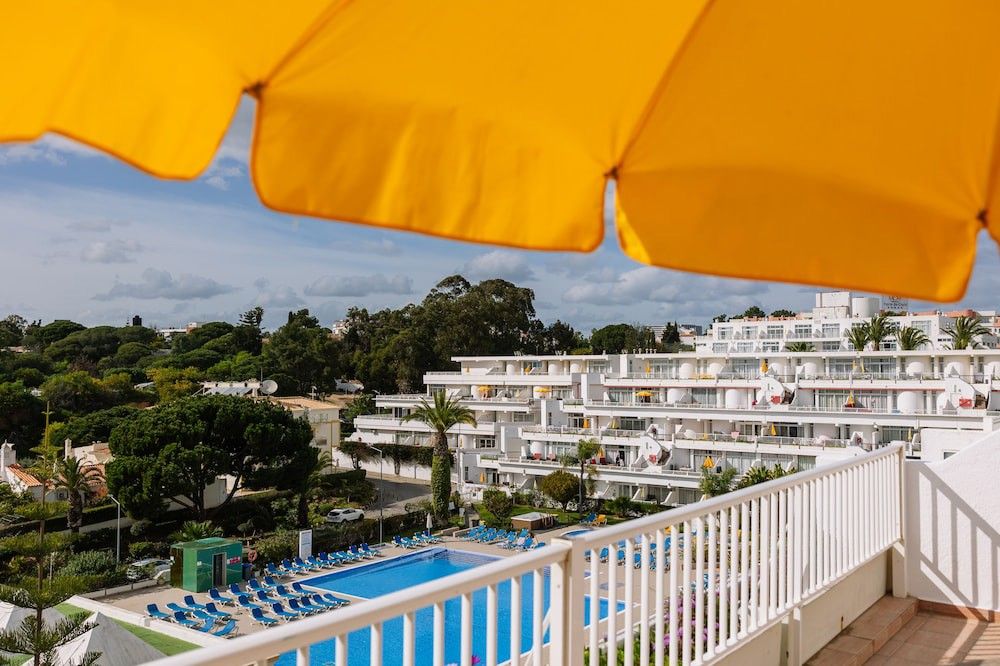 Utsikt från en balkong med vy över en resort med vita byggnader, en pool och träd, inramad av ett gult parasoll.