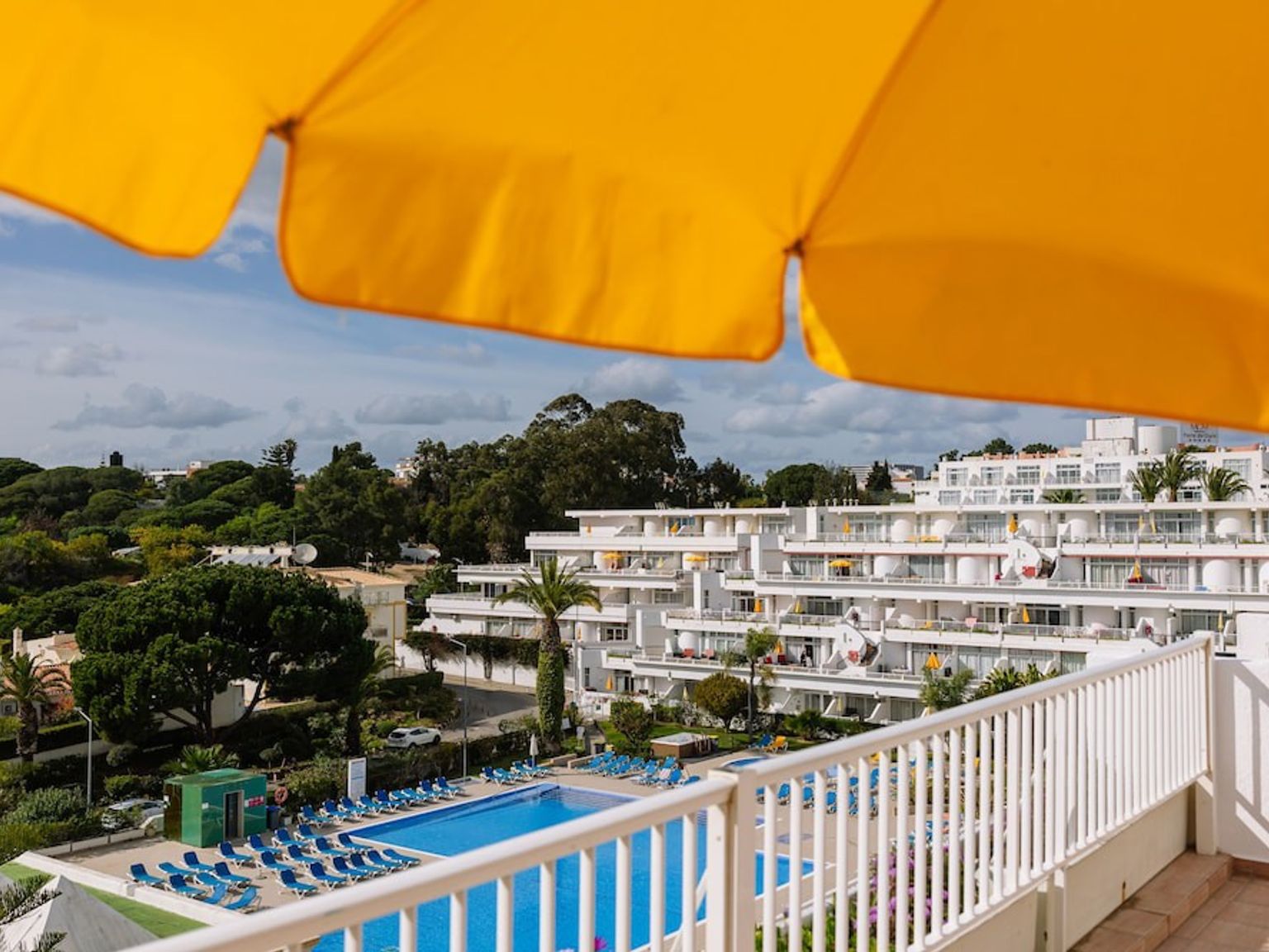 Utsikt från en balkong med vy över en resort med vita byggnader, en pool och träd, inramad av ett gult parasoll.