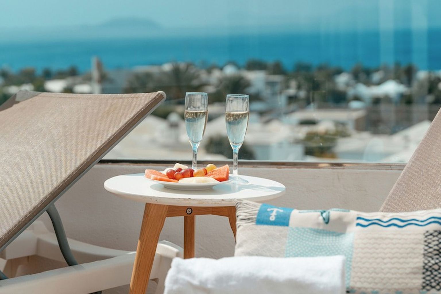 Balkong med solsenger, to glass champagne, en frukttallerken og havutsikt.