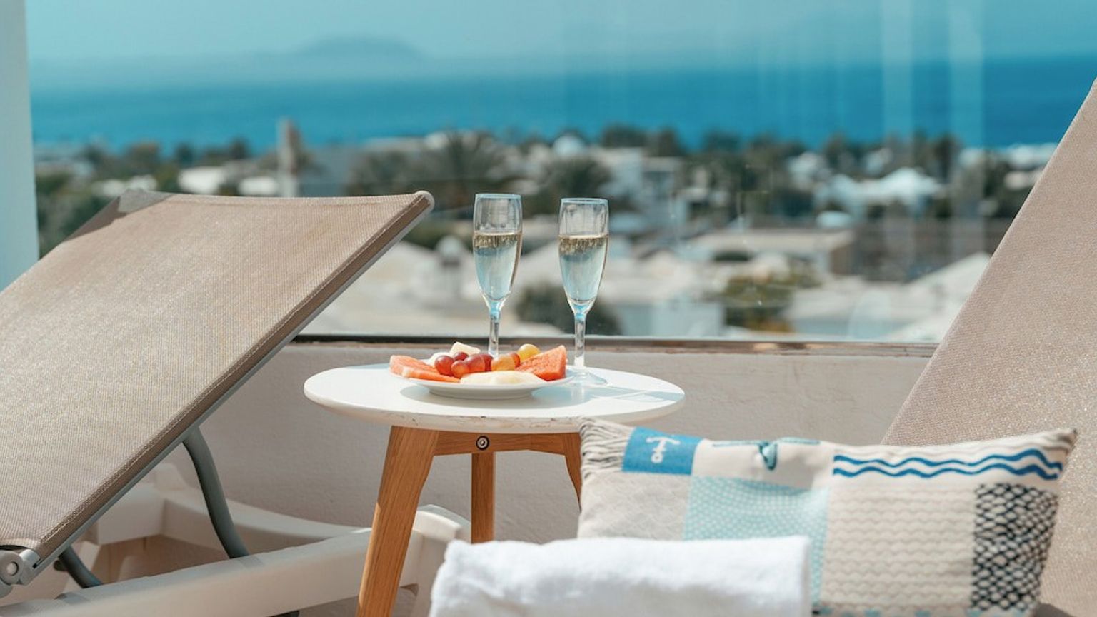 Balkong med solsenger, to glass champagne, en frukttallerken og havutsikt.