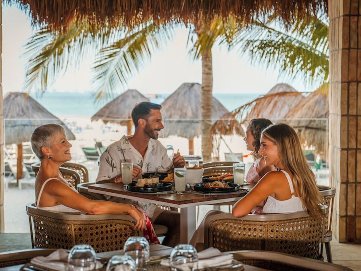 Fyra leende personer som äter på en utomhusrestaurang med utsikt över en tropisk strand med palmer.