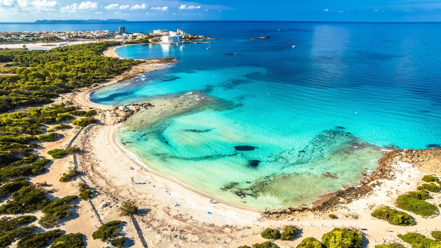 Luftfoto av en turkis bukt, sandstrand og grønne trær med en fjern kystby. Es Trenc, Mallorca