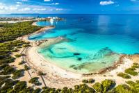 Luchtfoto van een turquoise baai, zandstrand en groene bomen met een verre kustplaats. Es Trenc, Mallorca