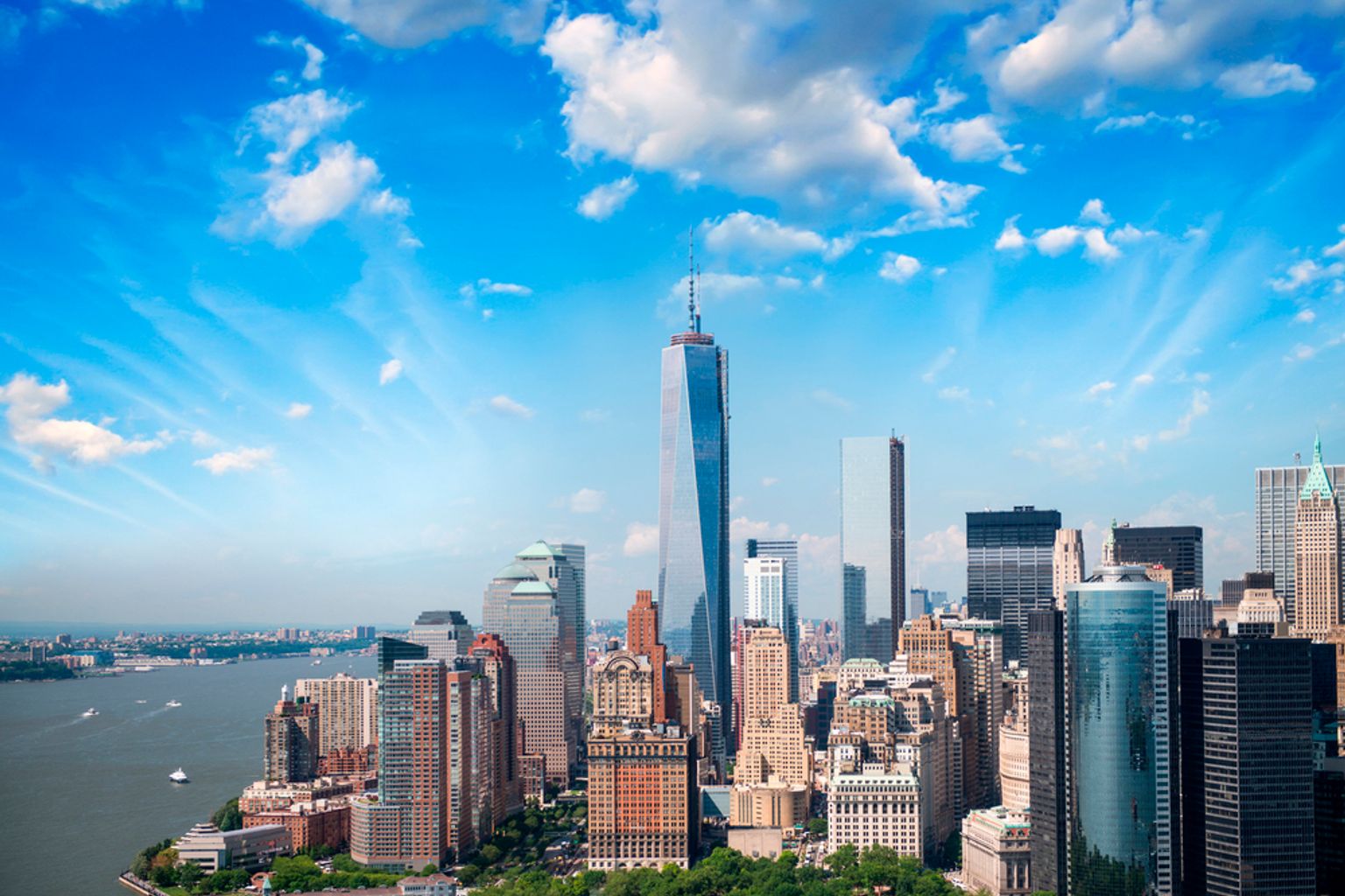Luftfoto af New Yorks skyline med One World Trade Center, en havnefront og en blå himmel med skyer.