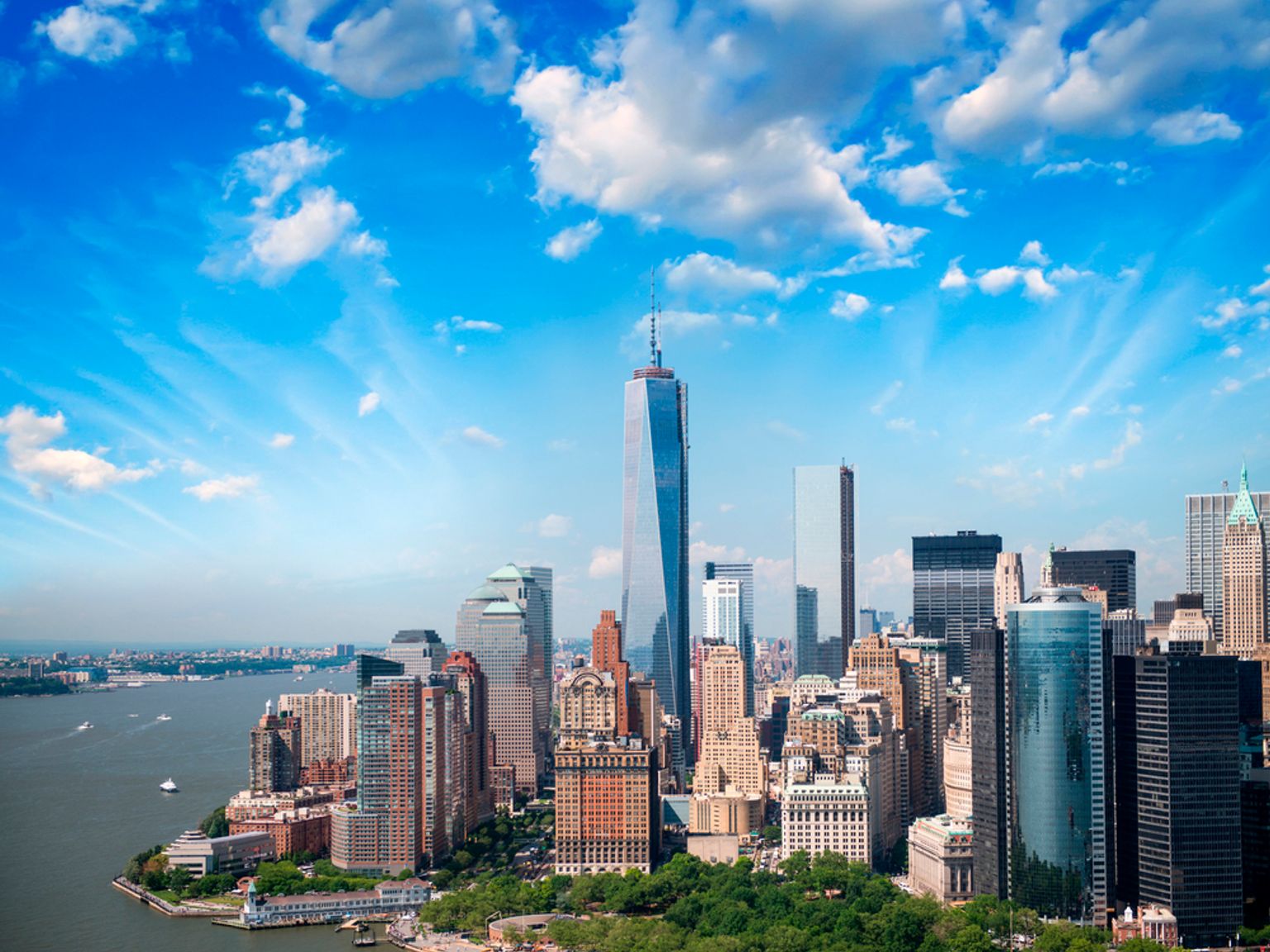 Luftfoto af New Yorks skyline med One World Trade Center, en havnefront og en blå himmel med skyer.