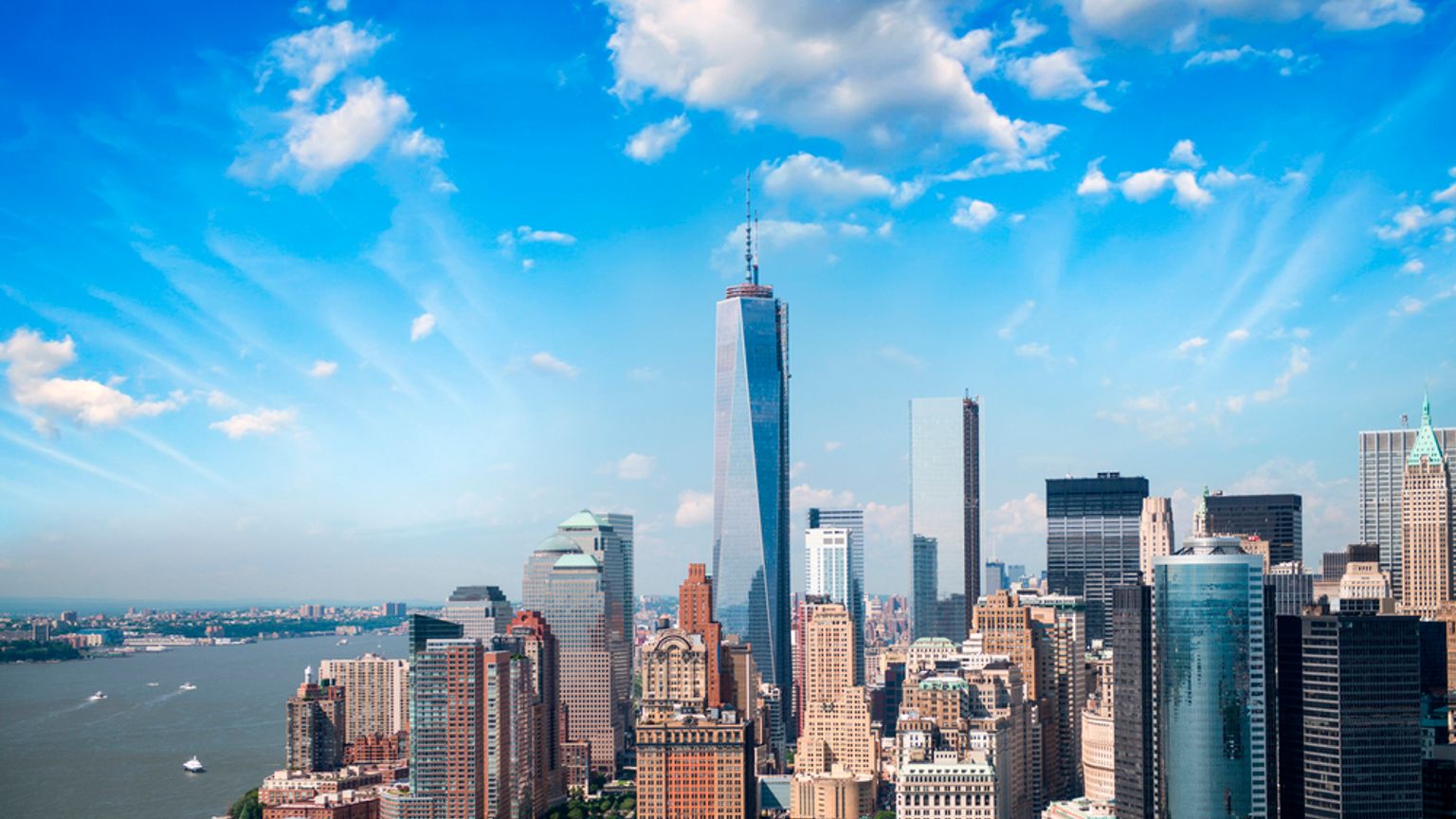 Luftfoto af New Yorks skyline med One World Trade Center, en havnefront og en blå himmel med skyer.
