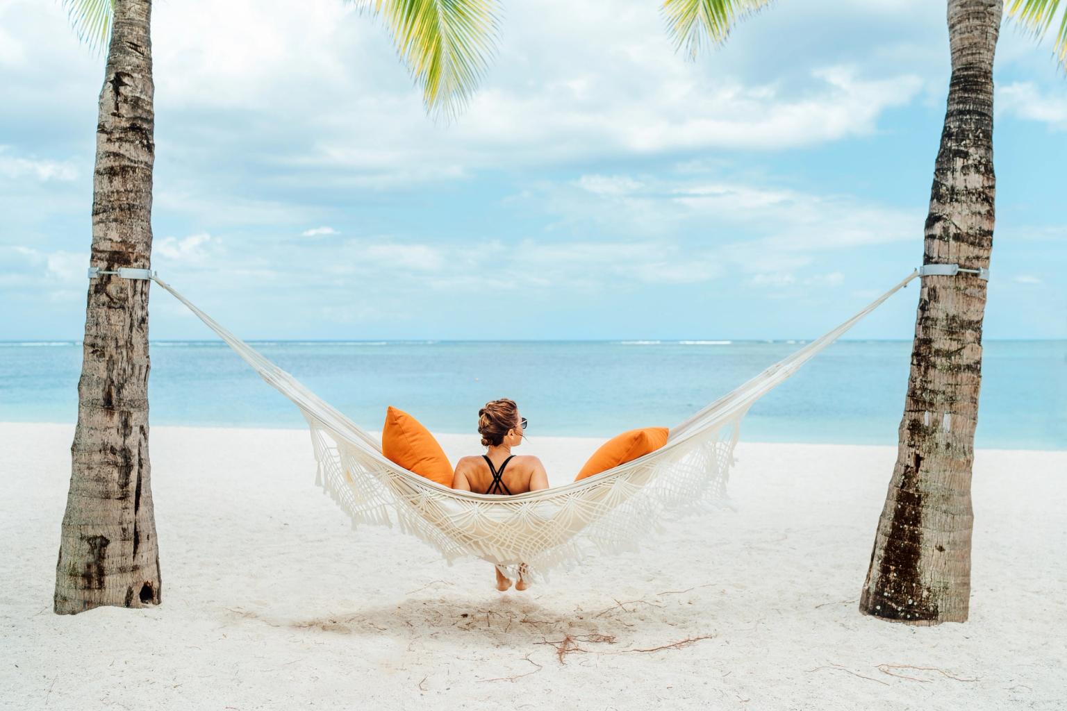 En person kopplar av i en hängmatta med orange kuddar mellan två palmer på en vit sandstrand, vänd mot havet.