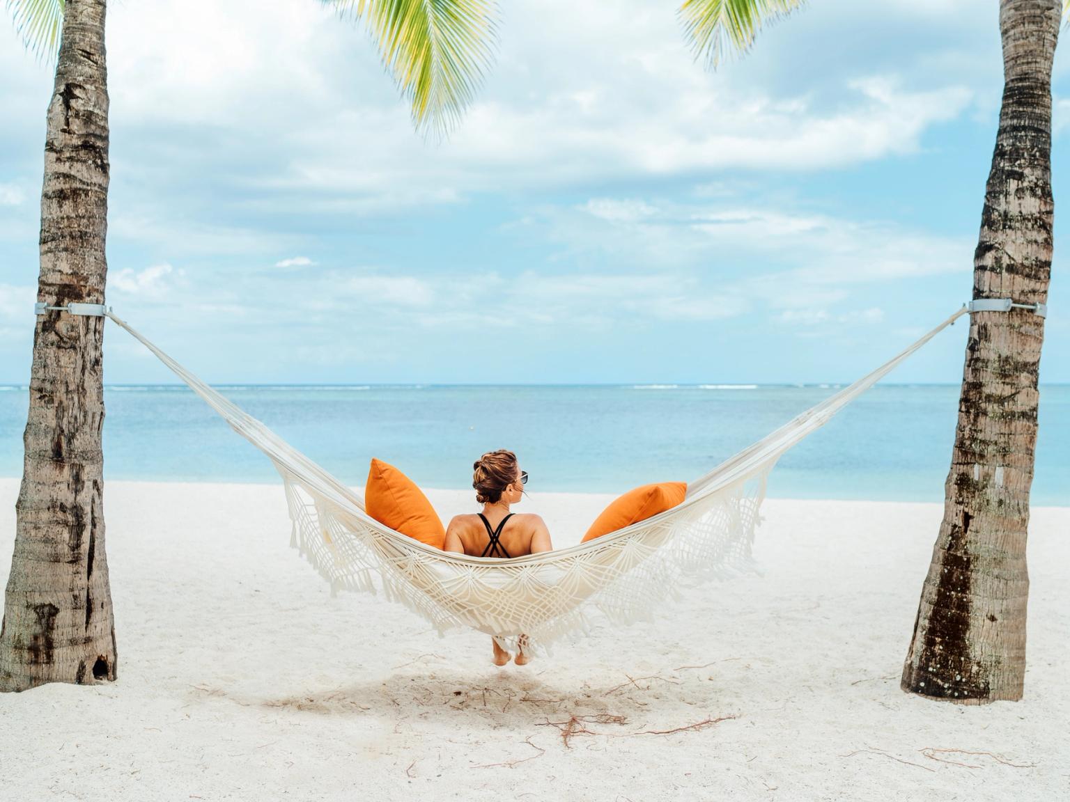 En person kopplar av i en hängmatta med orange kuddar mellan två palmer på en vit sandstrand, vänd mot havet.