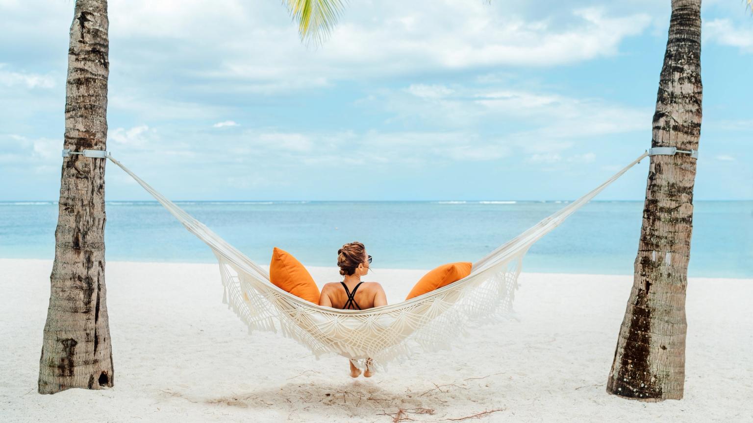 En person kopplar av i en hängmatta med orange kuddar mellan två palmer på en vit sandstrand, vänd mot havet.