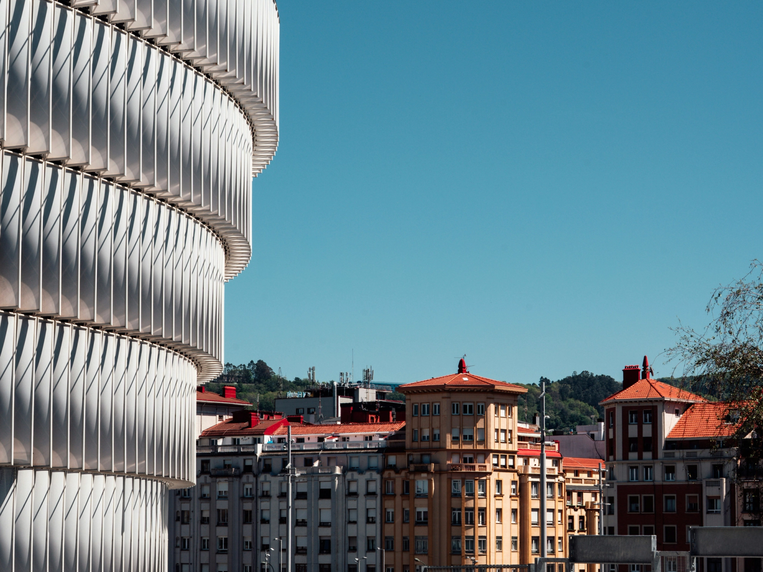 En modern, böjd vit byggnad med en ribbad fasad till vänster, i kontrast med traditionella byggnader under en klarblå himmel, och två män som står i förgrunden.