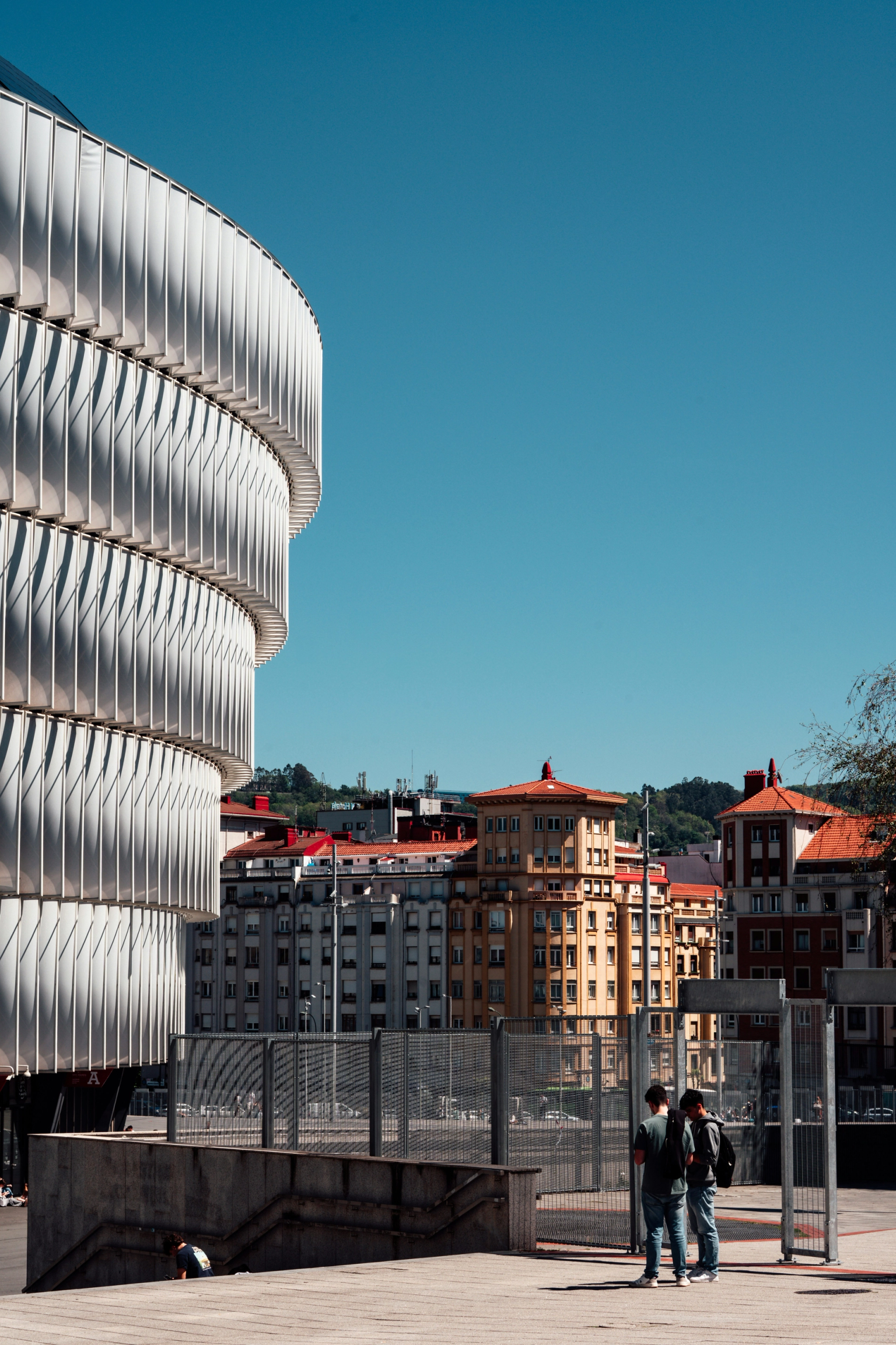 En modern, böjd vit byggnad med en ribbad fasad till vänster, i kontrast med traditionella byggnader under en klarblå himmel, och två män som står i förgrunden.