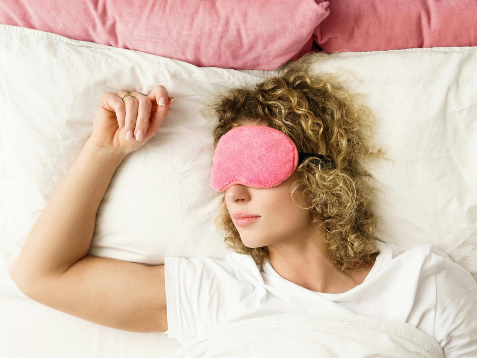 en kvinna sover i sängen med en rosa sovmask på ögonen.