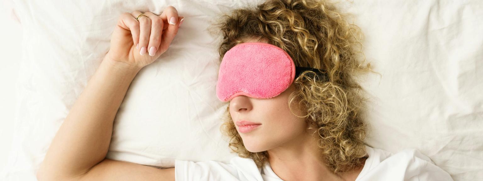 en kvinna sover i sängen med en rosa sovmask på ögonen.