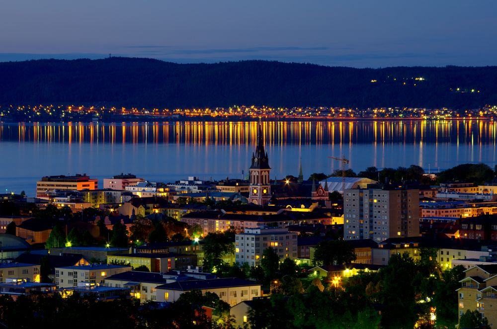 Nattutsikt over en by med en fremtredende kirkespir, lys som reflekteres i en stor vannflate, og en mørk fjellkjede i det fjerne.