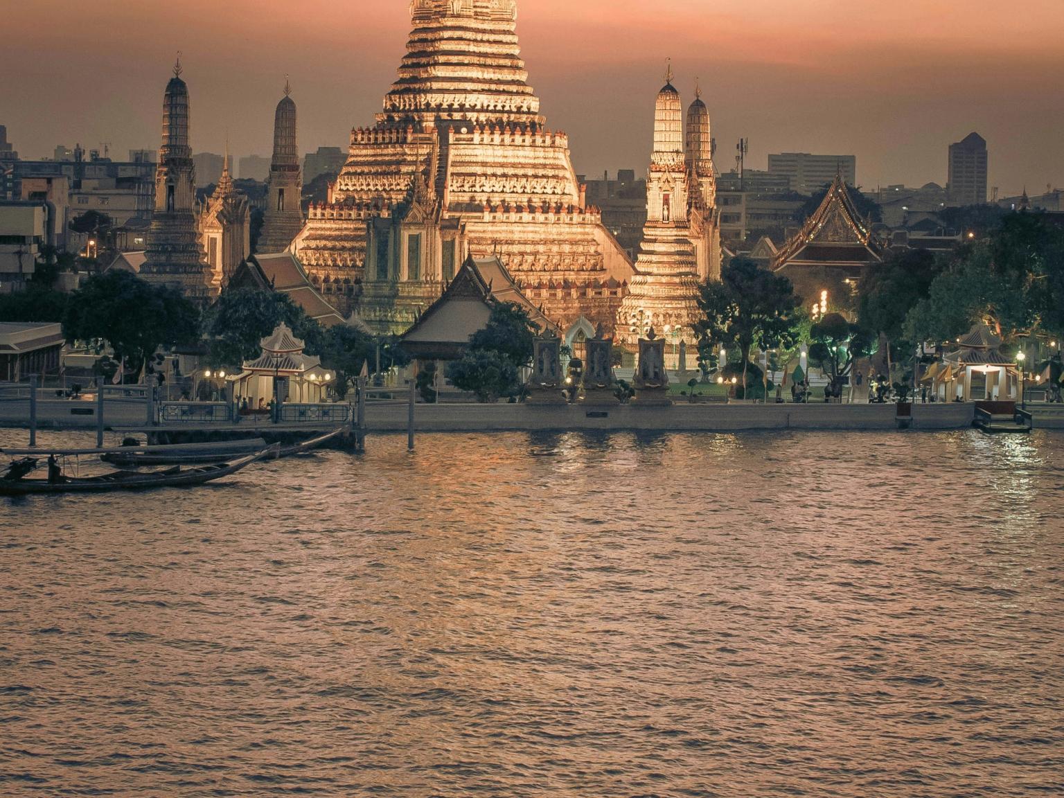 Oplyst Wat Arun-tempel over en flod ved solnedgang, med en langhalebåd. Bangkok
