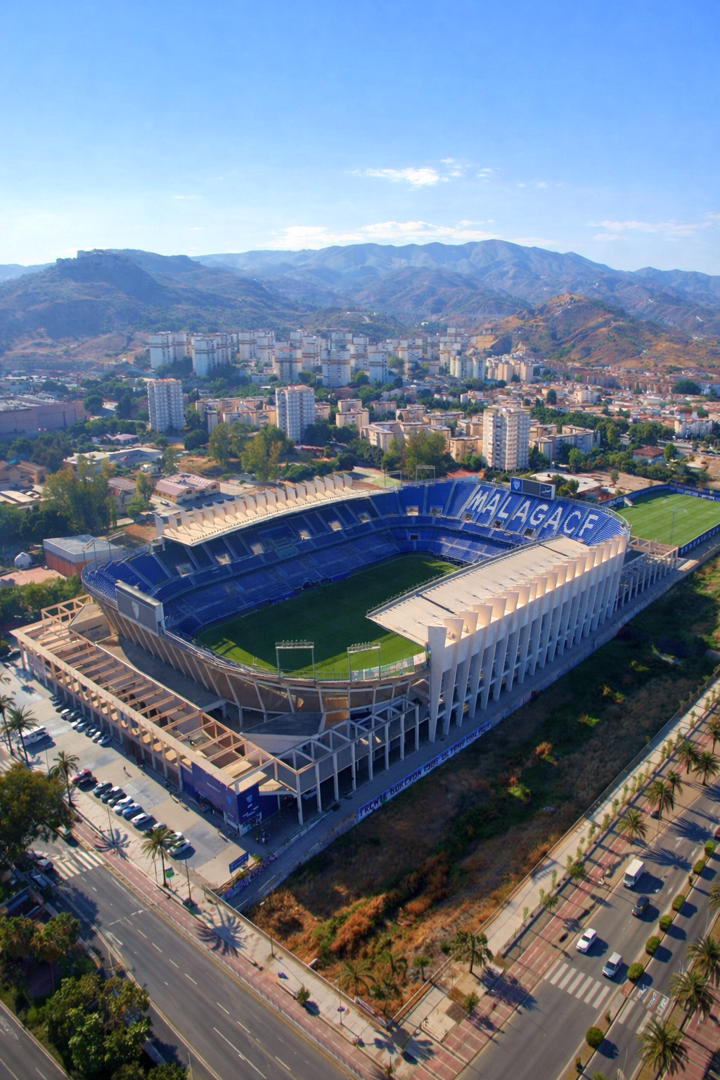 Aerial view of a football stadium with "MALAGACF" visible on blue seats, surrounded by a city and mountains under a clear sky.