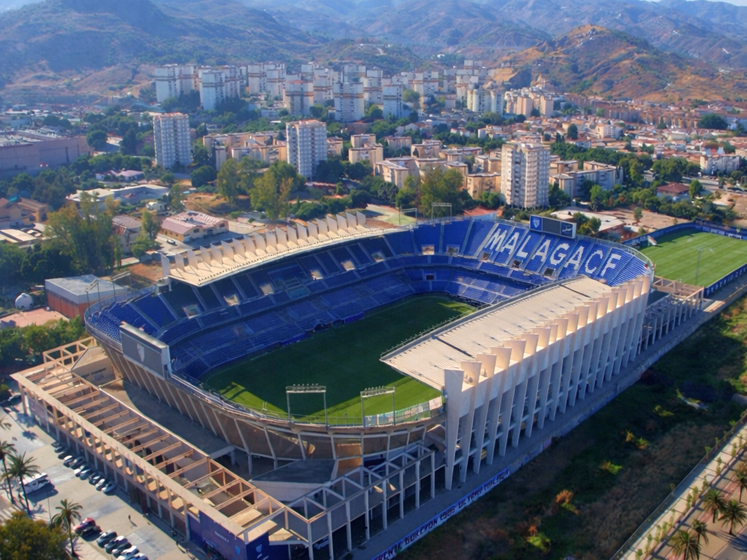 Aerial view of a football stadium with "MALAGACF" visible on blue seats, surrounded by a city and mountains under a clear sky.
