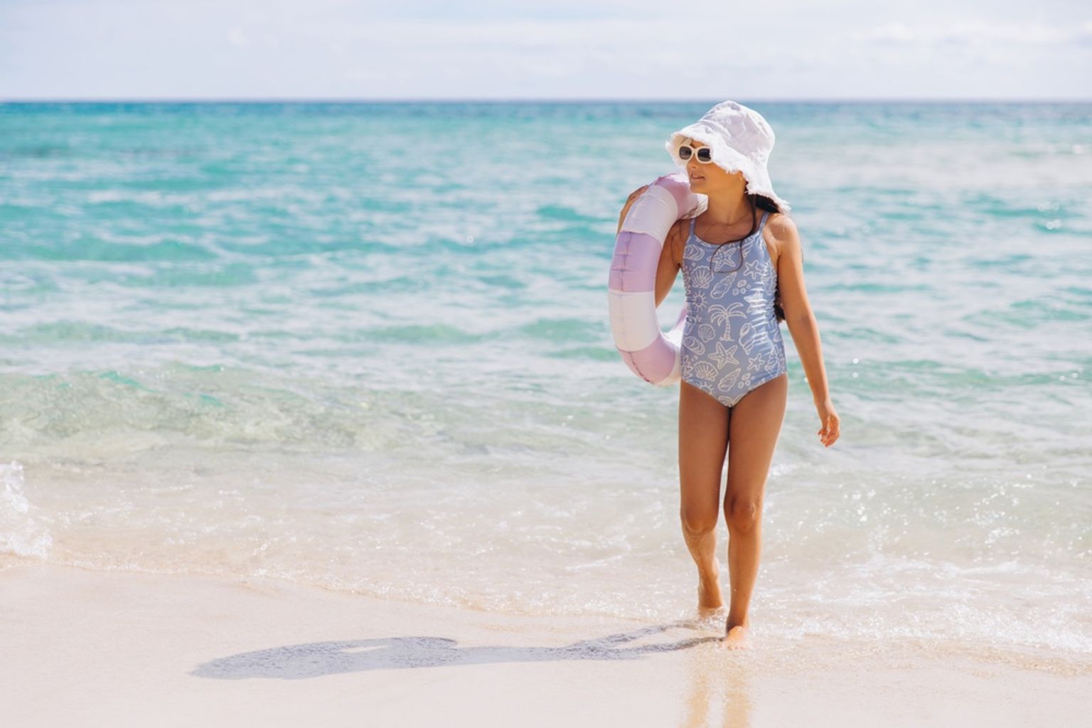 Ein junges Mädchen im Badeanzug, mit Hut und Sonnenbrille, geht an einem Sandstrand und trägt einen Schwimmring.