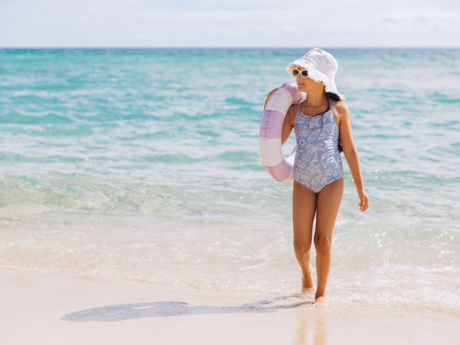 Młoda dziewczynka w stroju kąpielowym, kapeluszu i okularach przeciwsłonecznych idzie po piaszczystej plaży, niosąc koło do pływania.