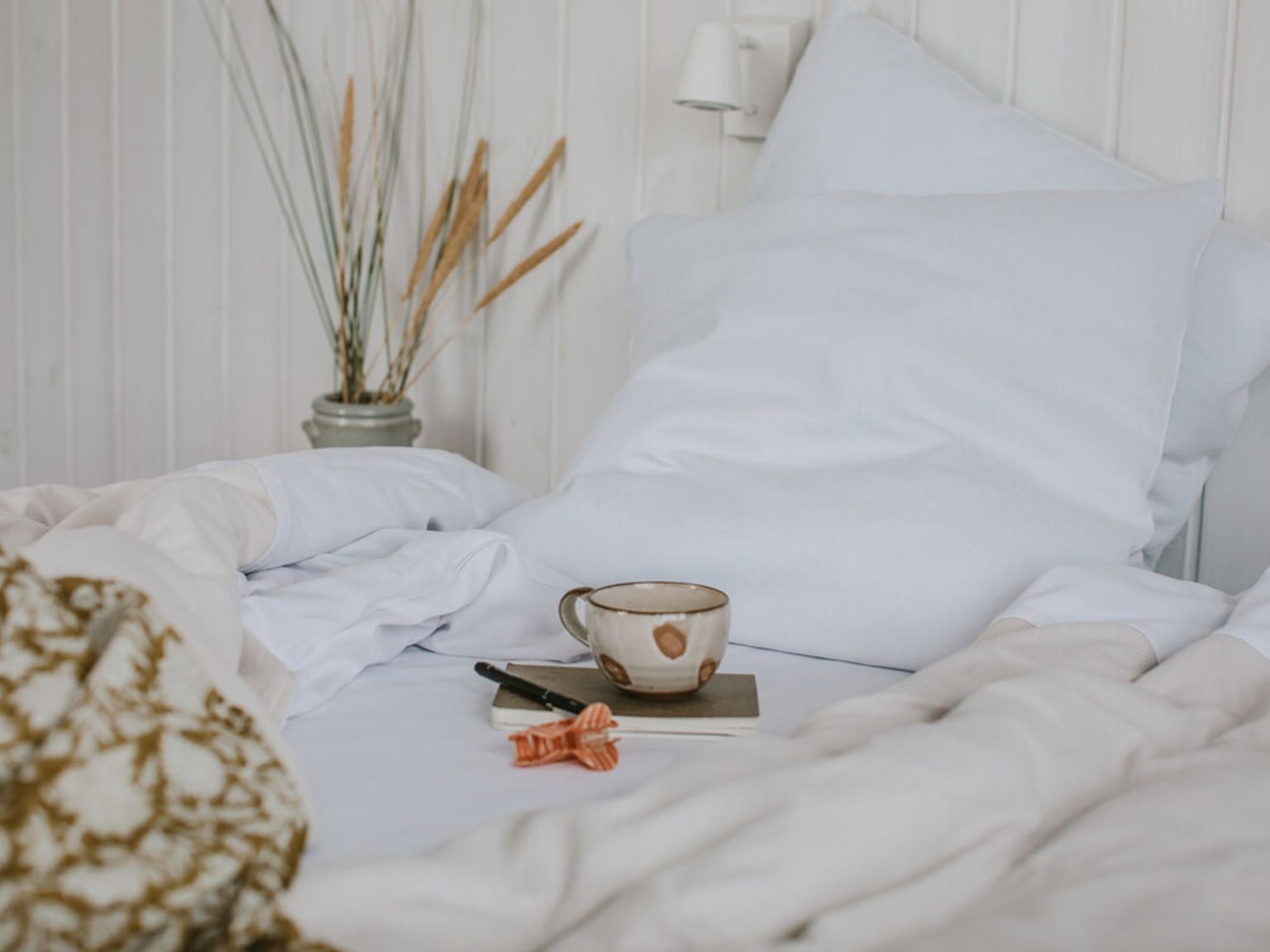 Hvit seng med en kopp, notatbok og tørket plante.