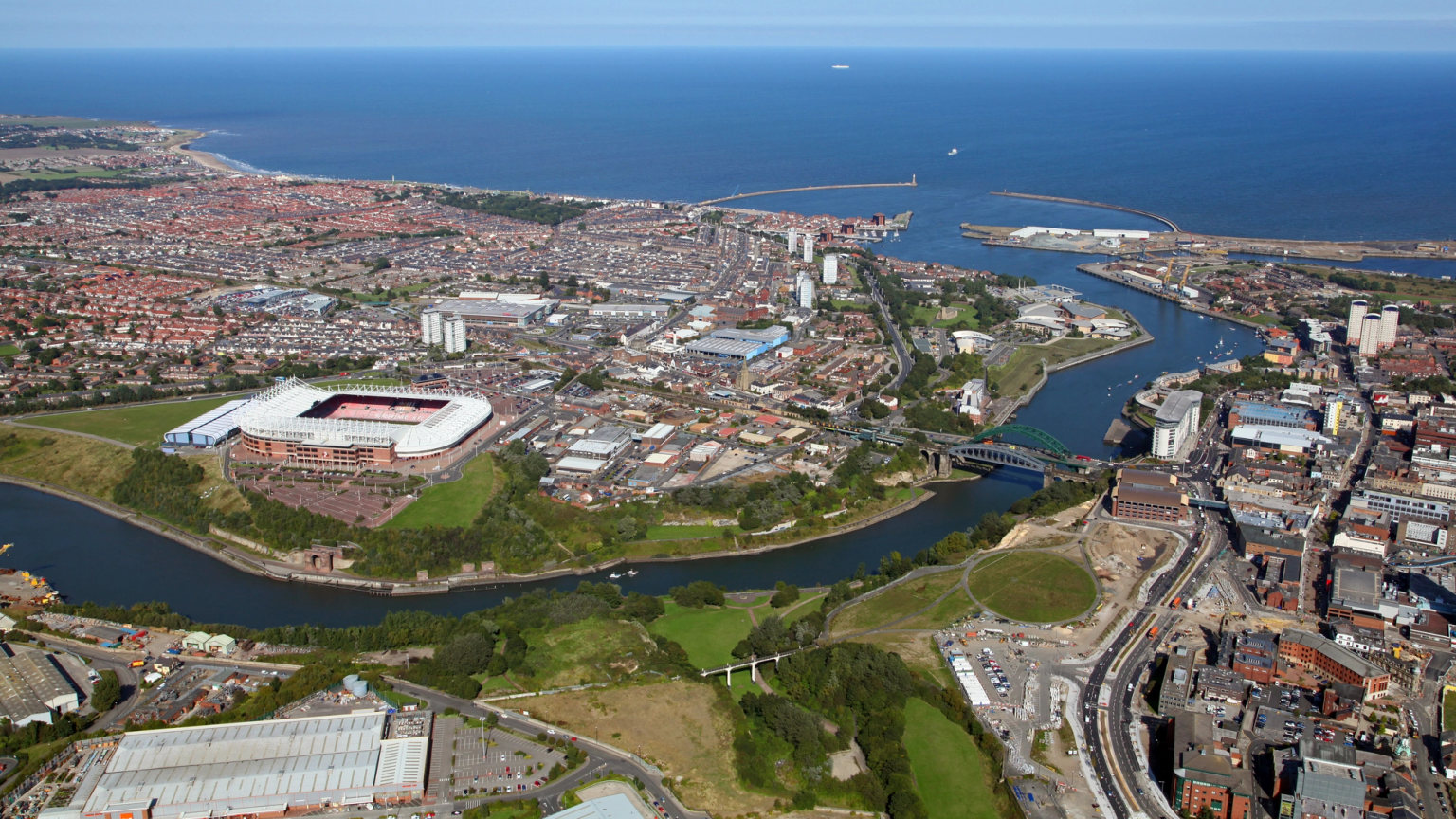 Aerial view of a city sprawling along a river and coastline, featuring a stadium and a port.