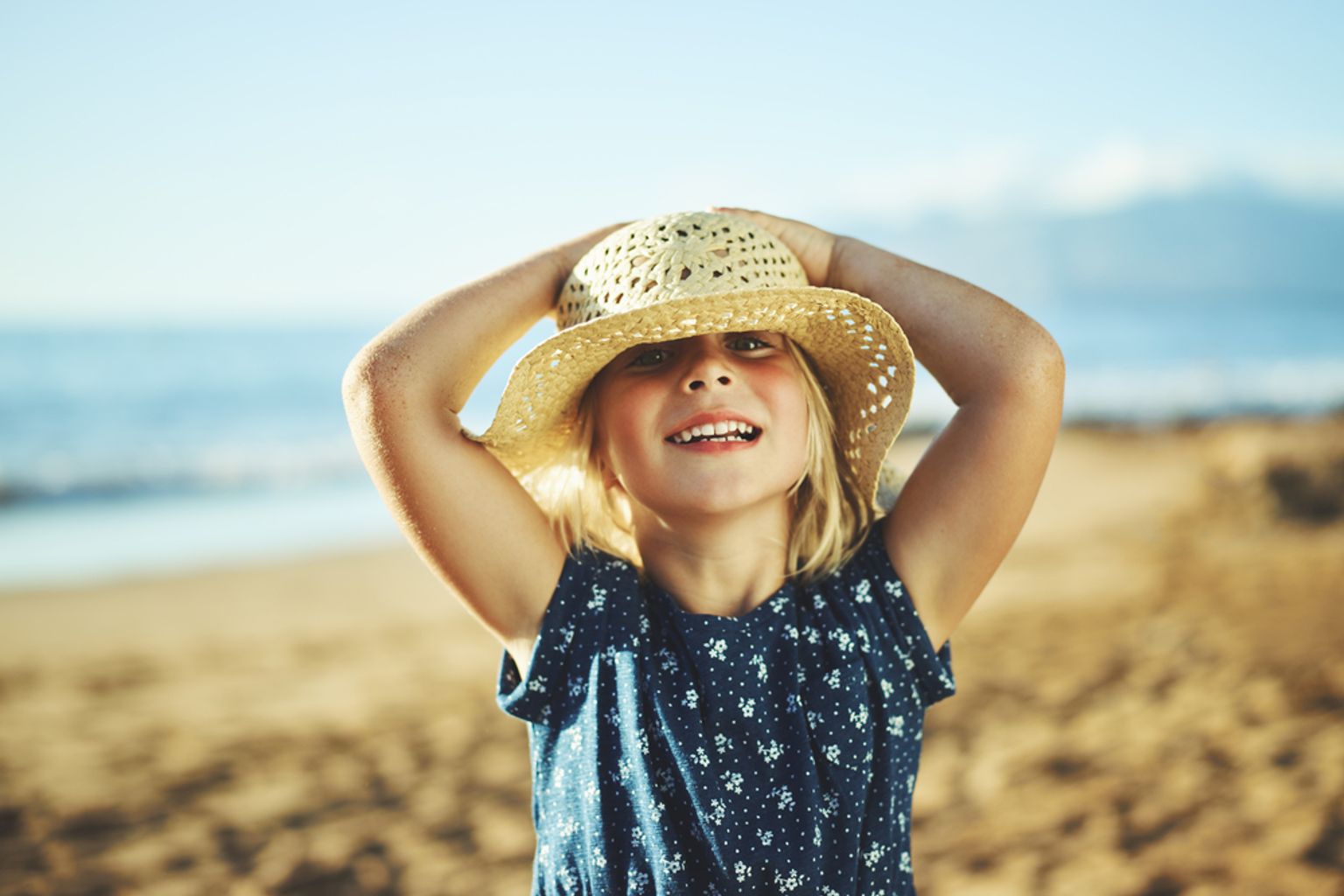 En blond pige med en stråhat smiler på en solrig strand.