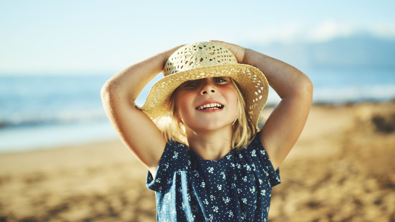 En blond pige med en stråhat smiler på en solrig strand.