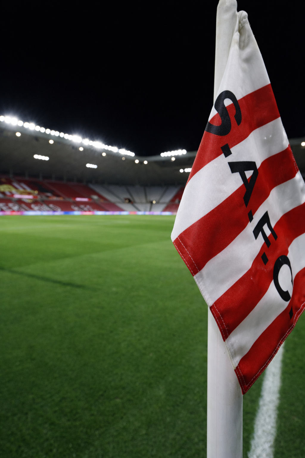 Red and white striped corner flag with "S.A.F.C." on a football pitch, with a floodlit stadium in the background.