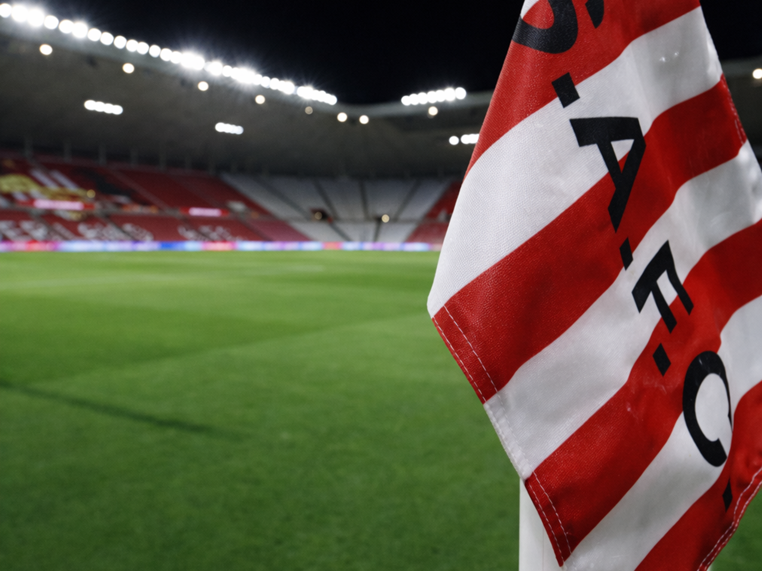 Red and white striped corner flag with "S.A.F.C." on a football pitch, with a floodlit stadium in the background.