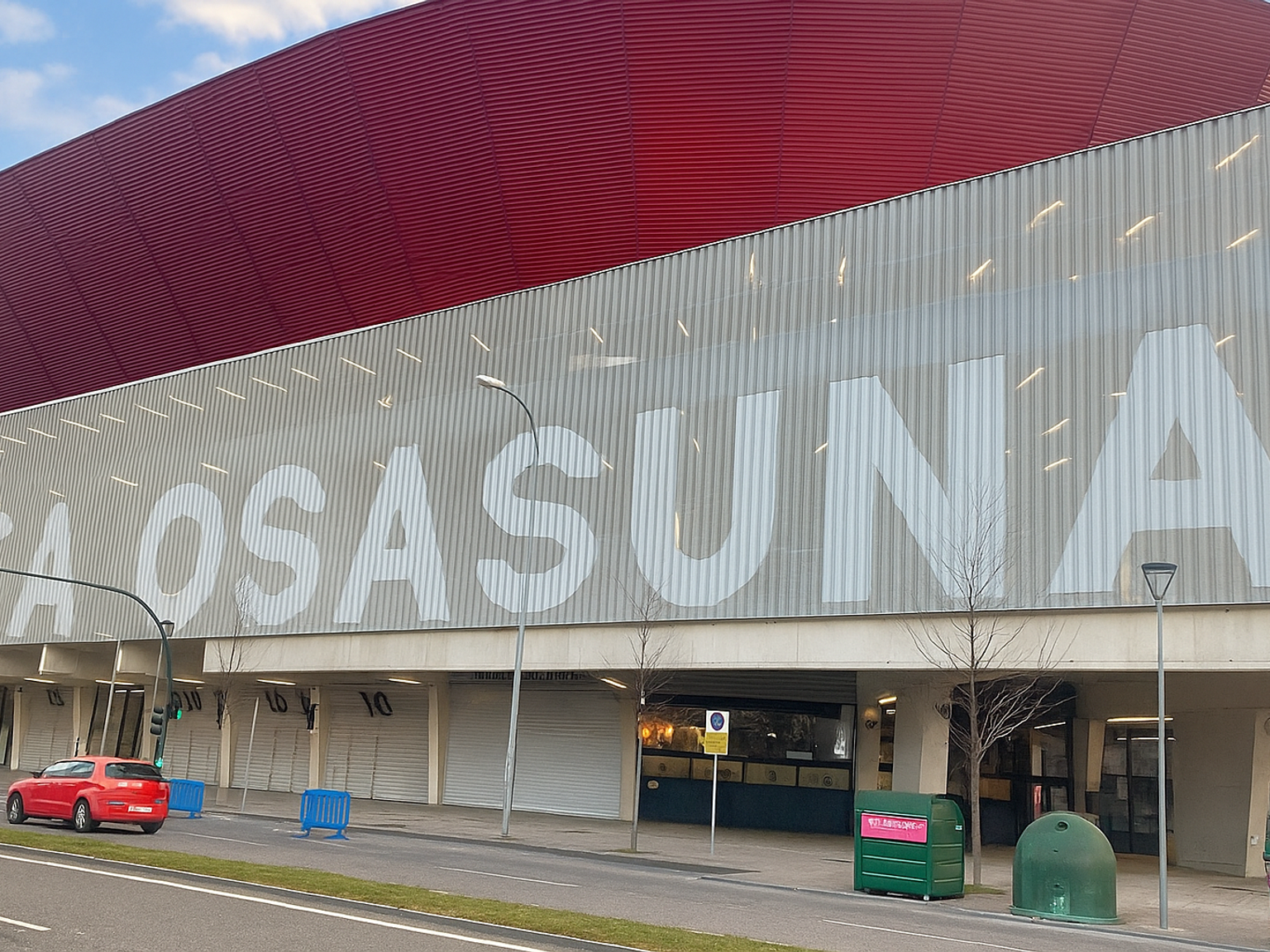 Een stadion met een rood dak en "CA OSASUNA" geschreven in grote witte letters op de grijze gevel, gezien vanaf een straat.