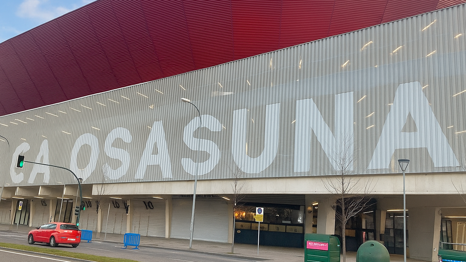 A stadium with a red roof and "CA OSASUNA" written in large white letters on its gray facade, seen from a street.