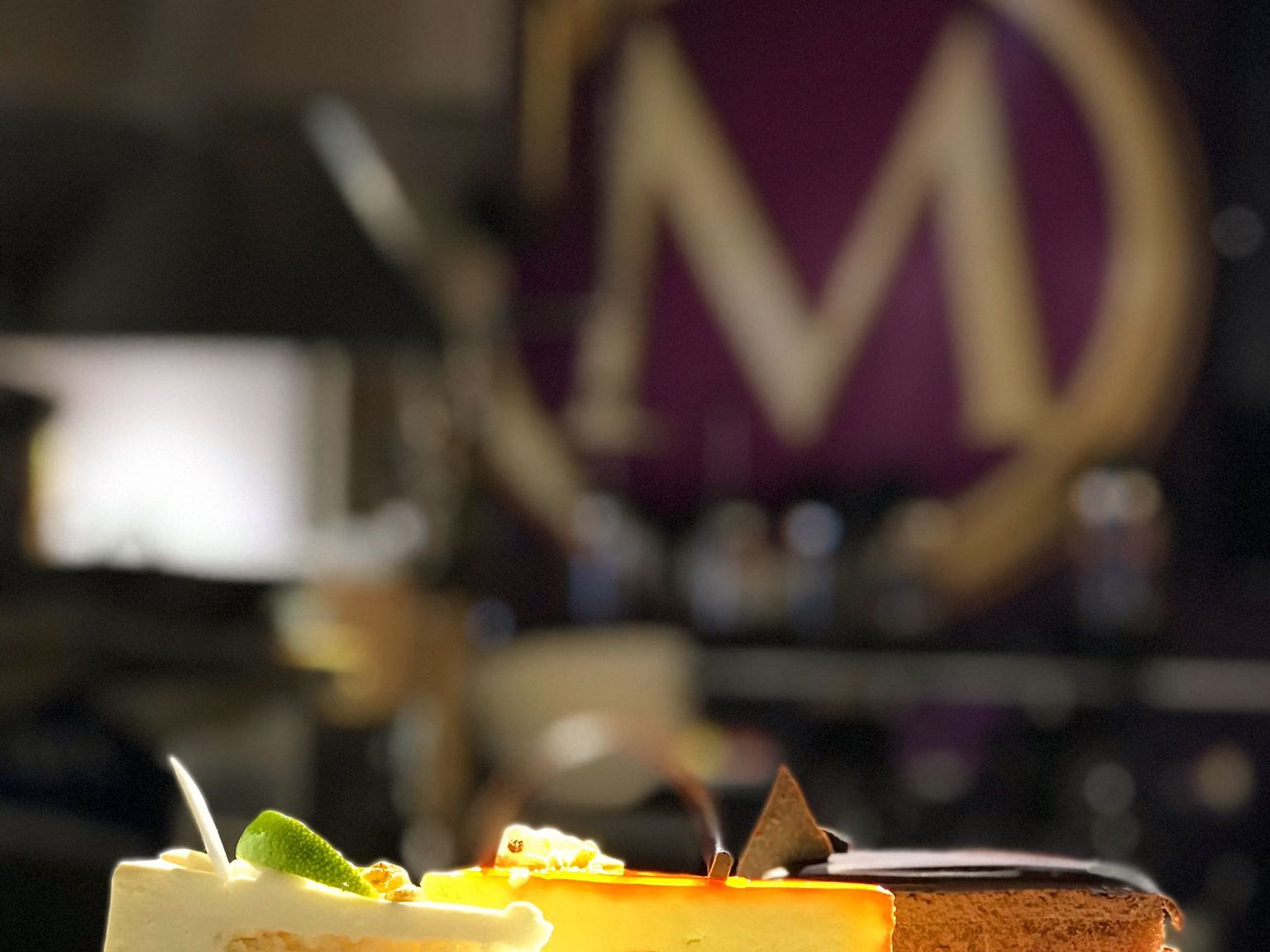 Kolme värikästä kakkupalaa—sitruuna, juustokakku ja suklaamousse—valkoisella lautasella, taustalla sumea violetti 'M'-logo.