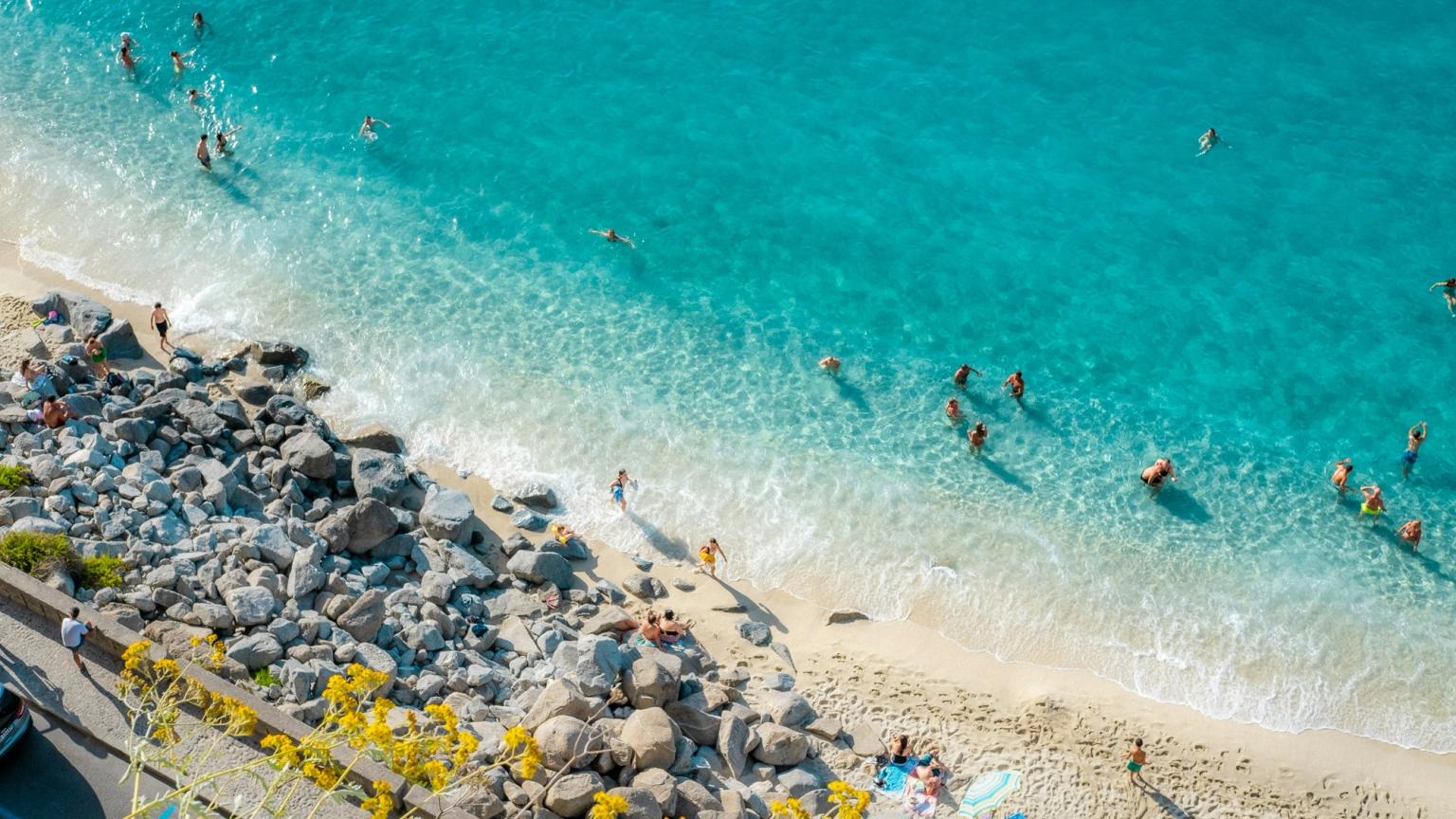 Aerial view of a lively beach with clear turquoise water, people swimming, and sunbathers. Calabria, Italy