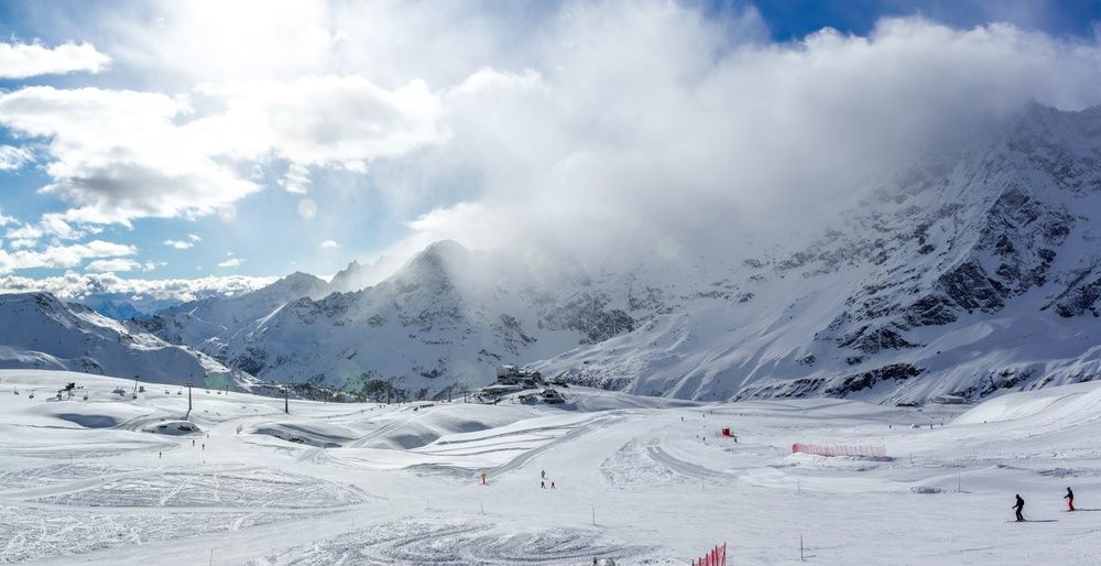Panoramautsikt over et snødekt skianlegg i fjellet med preparerte løyper og skyer som delvis skjuler fjerne fjelltopper.