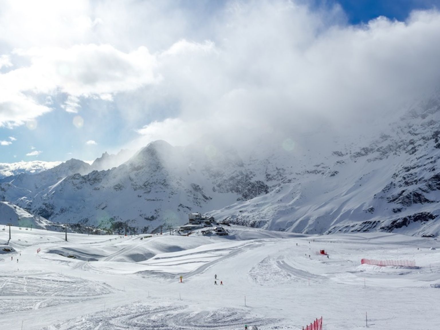 Panoramautsikt over et snødekt skianlegg i fjellet med preparerte løyper og skyer som delvis skjuler fjerne fjelltopper.