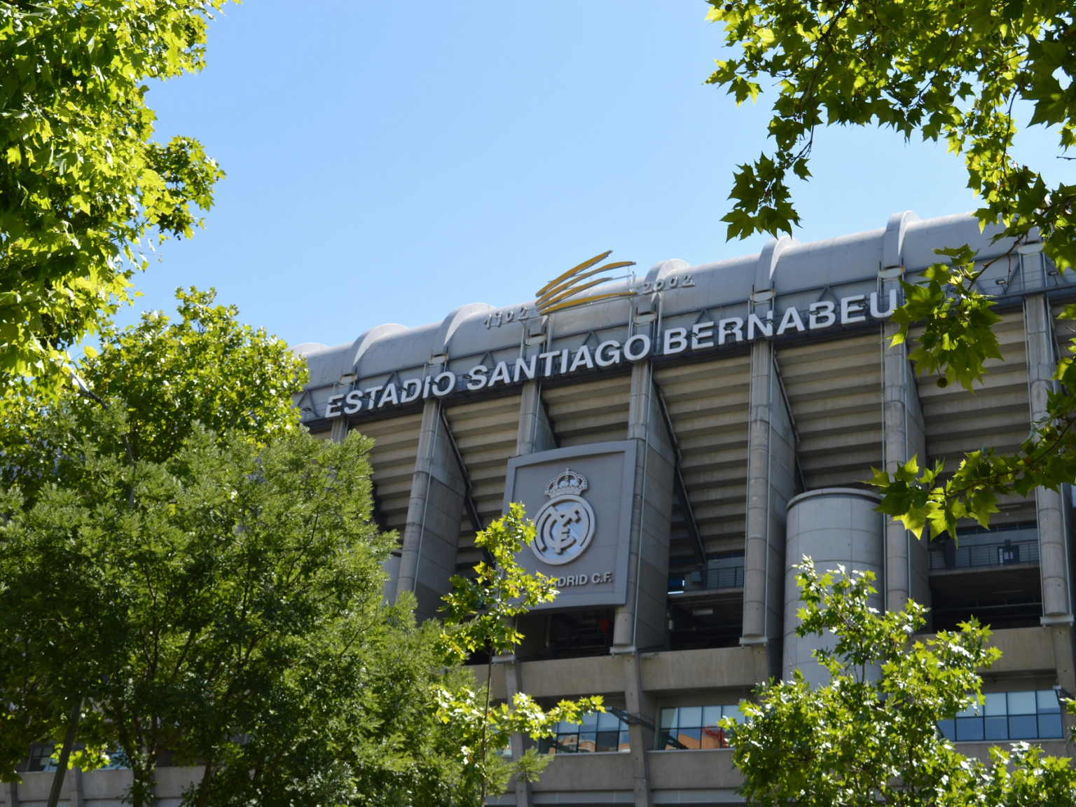 A Santiago Bernabéu Stadion külseje a Real Madrid címerével, részben zöld fák által eltakart.