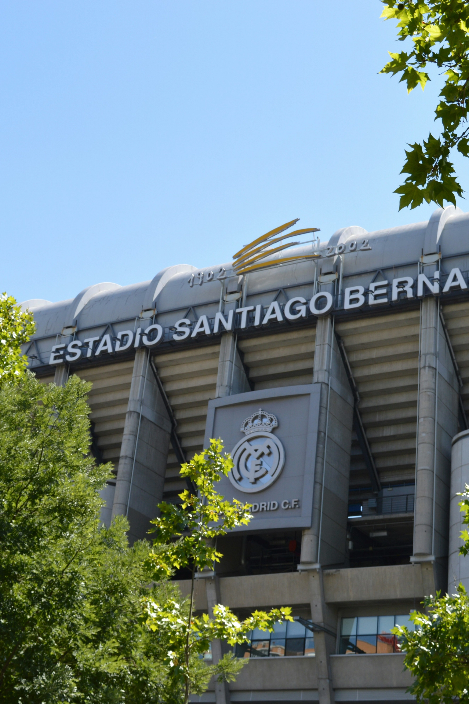A Santiago Bernabéu Stadion külseje a Real Madrid címerével, részben zöld fák által eltakart.