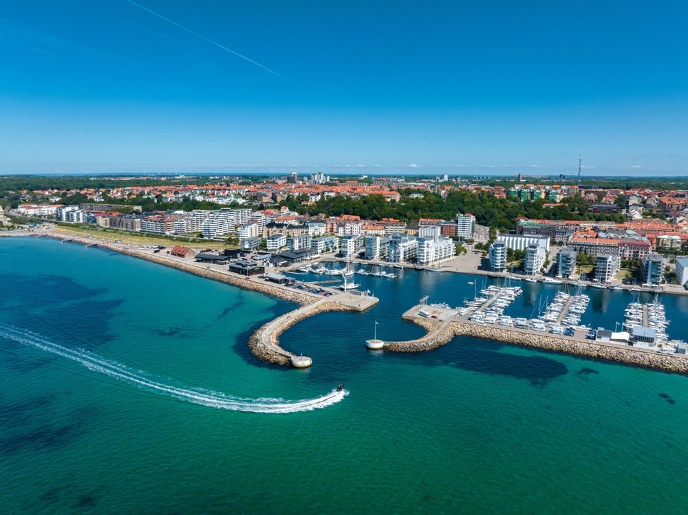 Luftfoto av en kystby med en sandstrand, en marina full av båter og en vannscooter på turkis vann.