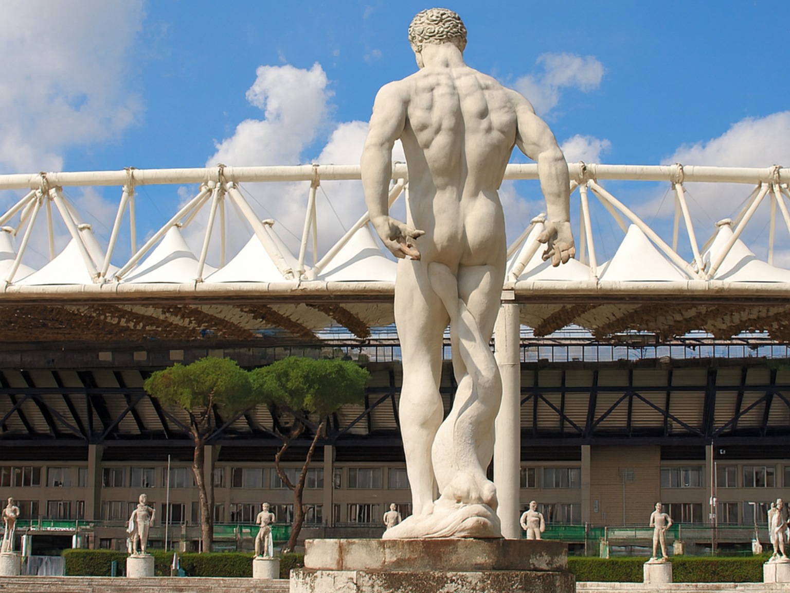 Bakifrån av en muskulös vit marmorskulptur på en piedestal, placerad mot en stadion med ett vitt tältat tak och en röd löparbana under en blå himmel.