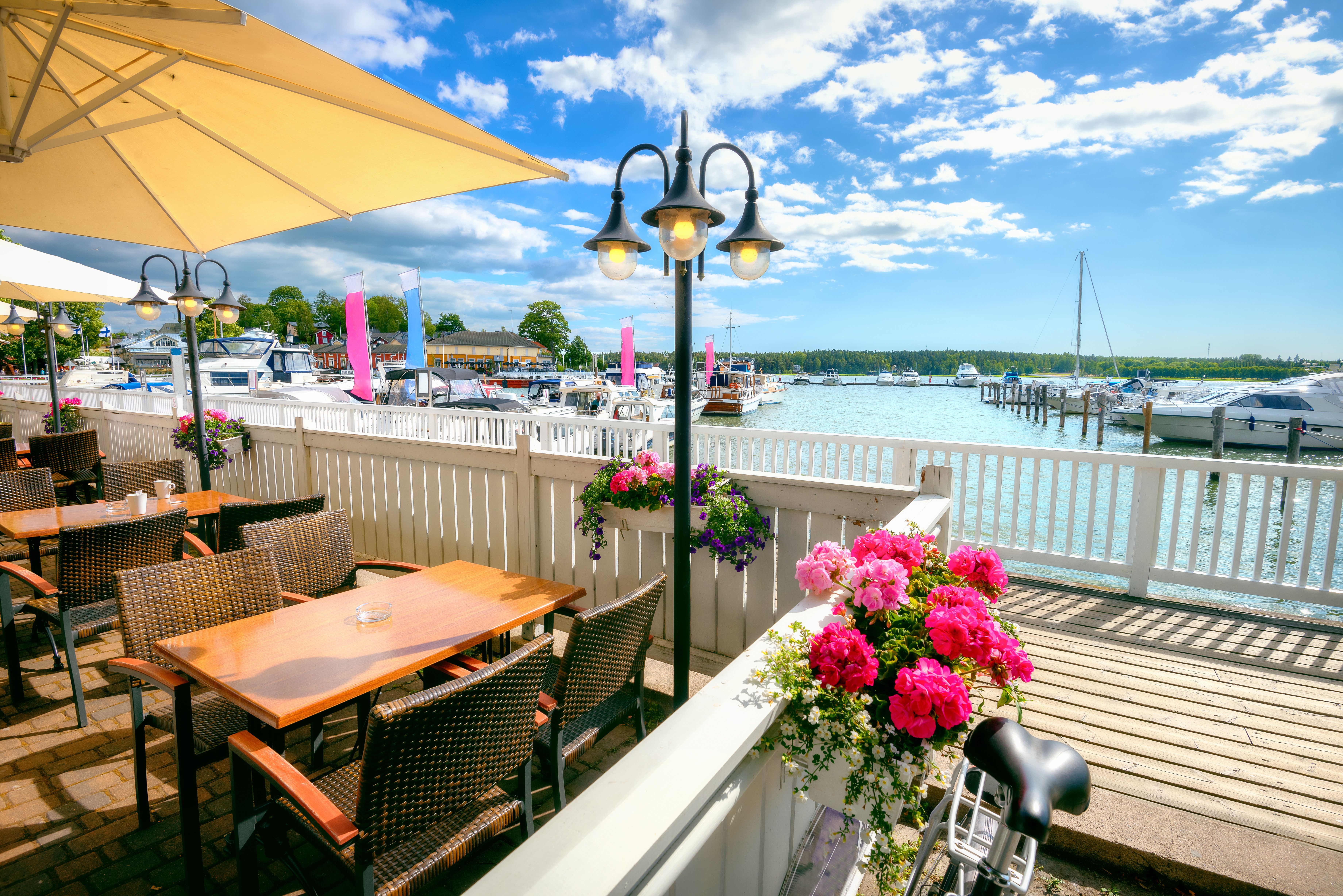 Solrig udendørs restaurantterrasse med lyserøde blomster med udsigt over en marina fyldt med både.