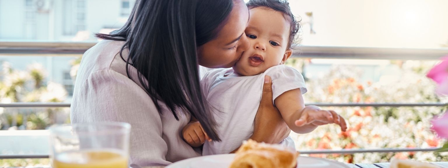 une femme tient un bébé tout en étant assise à une table avec une assiette de nourriture.