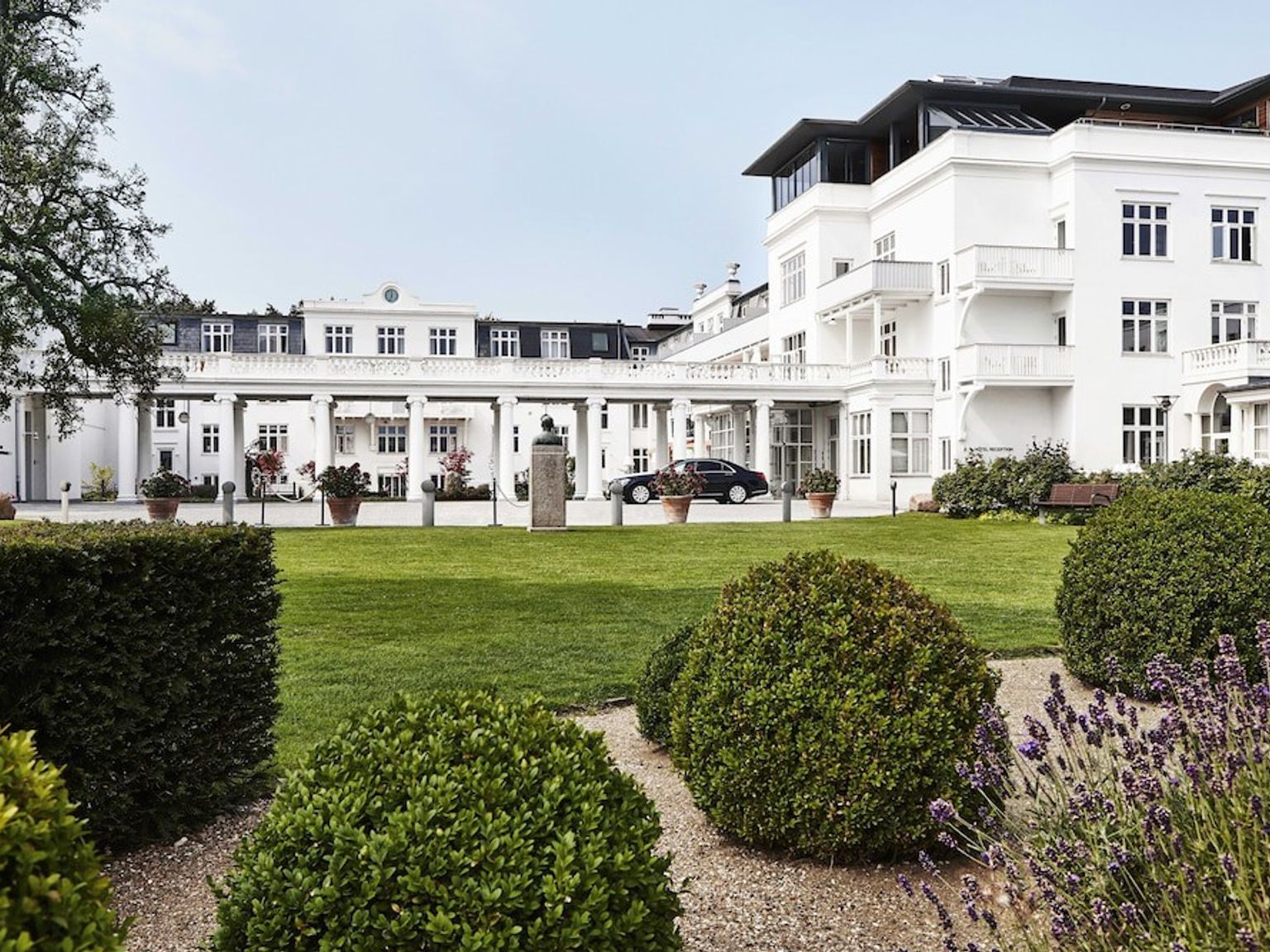 Elegant hvid bygning med en søjleprydet indgang og altaner, set fra en velplejet have med en grøn græsplæne.