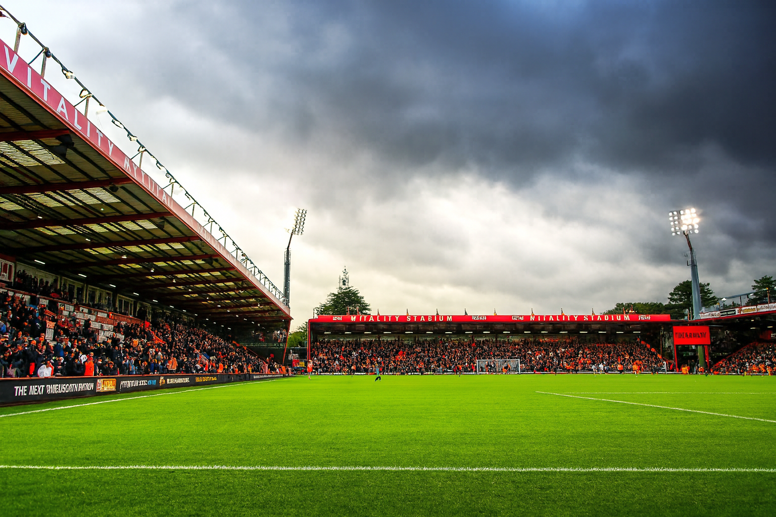A football stadium with a green pitch, packed stands, and bright floodlights under a cloudy sky.