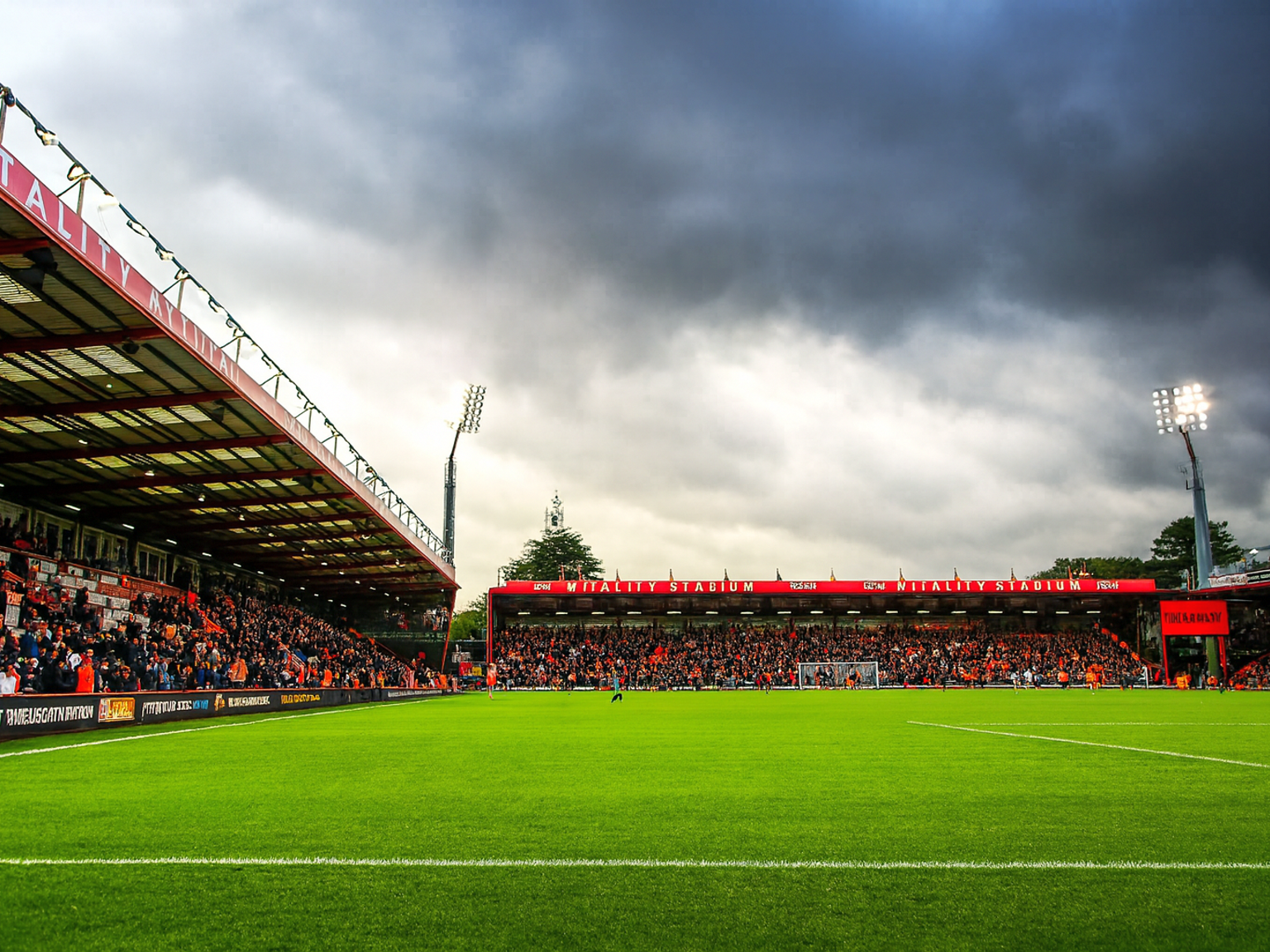 Ein Fußballstadion mit einem grünen Spielfeld, vollen Tribünen und hellen Flutlichtstrahlern unter einem bewölkten Himmel.