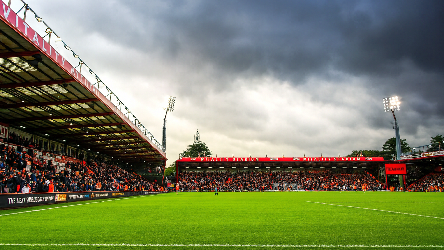 A football stadium with a green pitch, packed stands, and bright floodlights under a cloudy sky.