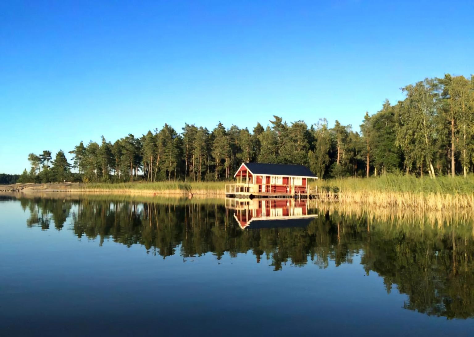 Rødt bådehus, der spejler sig i roligt søvand, omgivet af en skov under en klar blå himmel.